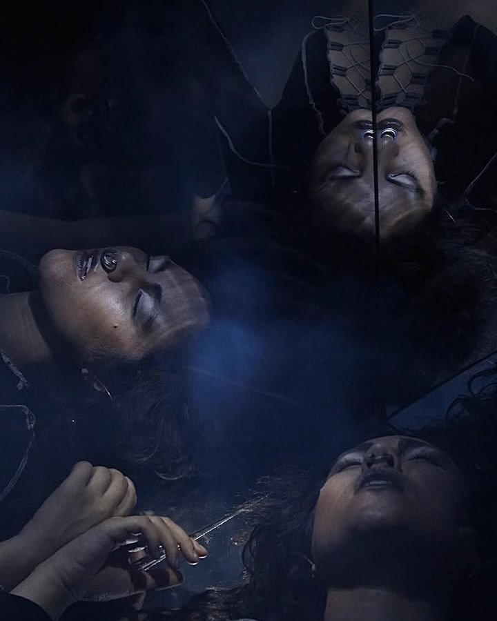 Female figure lying on the ground in a dark room with mirrors reflecting their face in multiple directions.